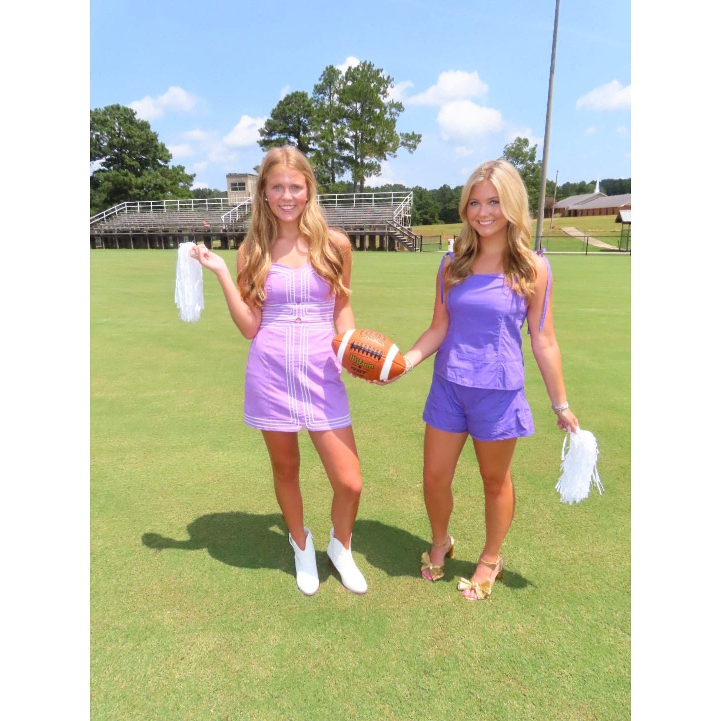 Two young women standing on a grassy sports field under a blue sky, wearing matching purple outfits. One holds a white pom-pom, and the other holds a white pom-pom while the woman on the left also holds an American football.
