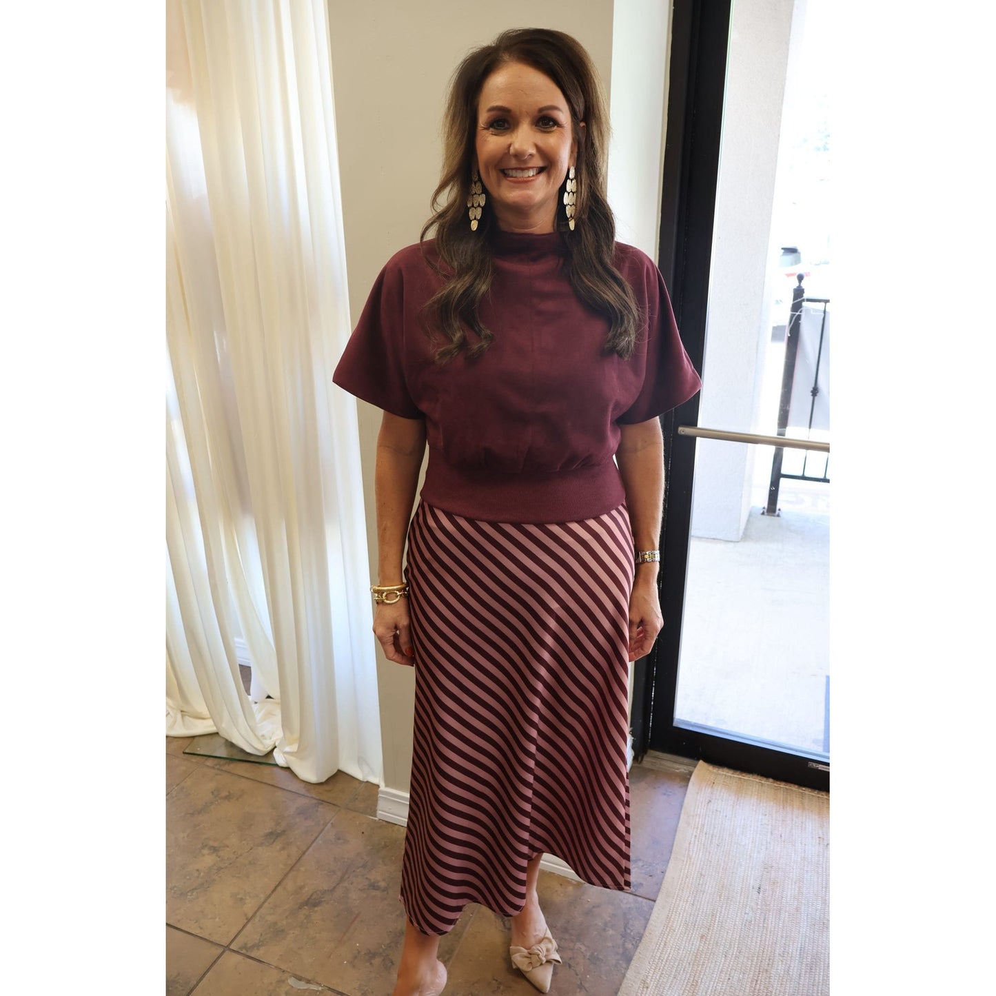 A woman modeling a two-piece outfit consisting of a short-sleeve mock neck top in burgundy and a matching long skirt with diagonal stripes in burgundy and white, standing in an indoor setting.