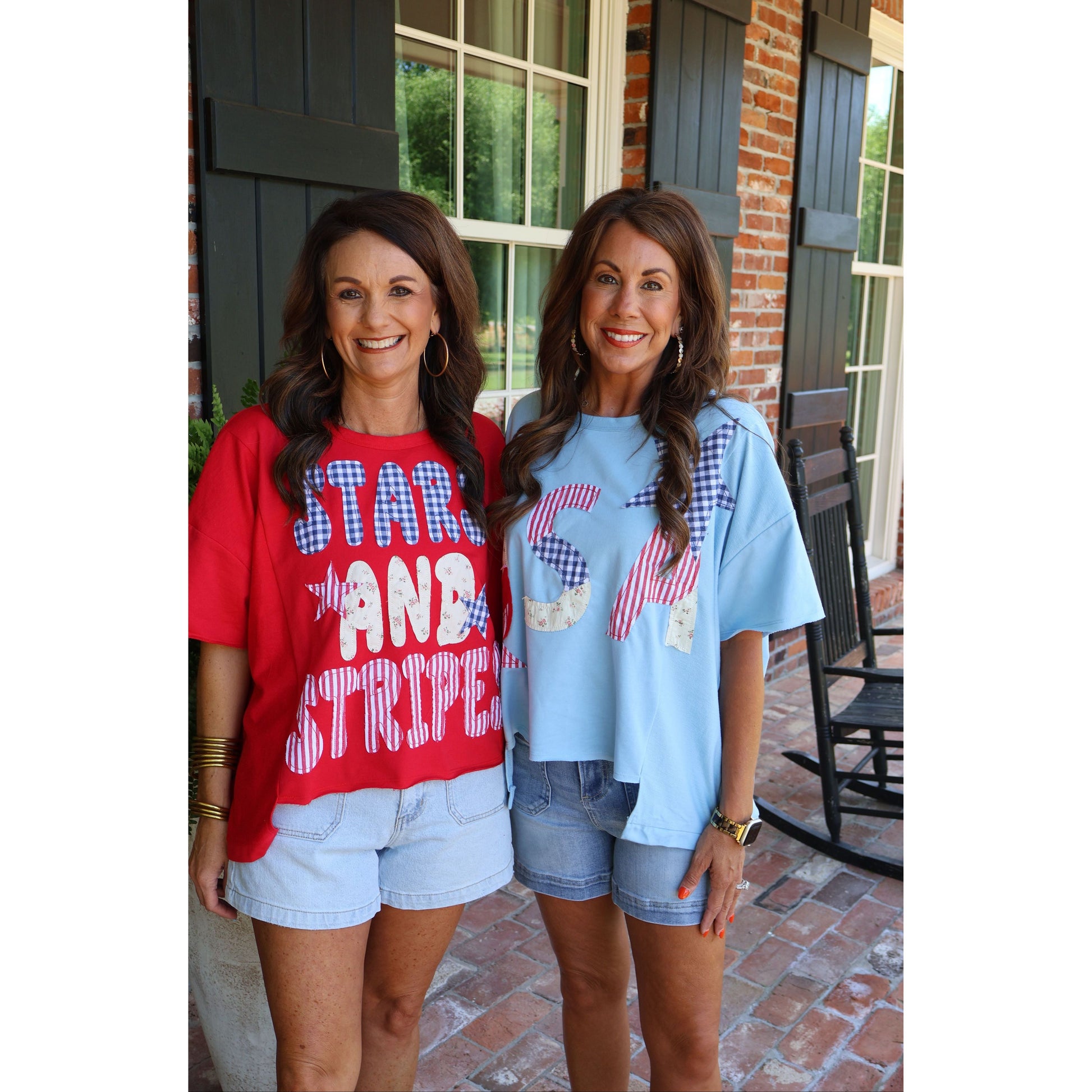 Two women standing outside a house, wearing red and blue t-shirts with text.