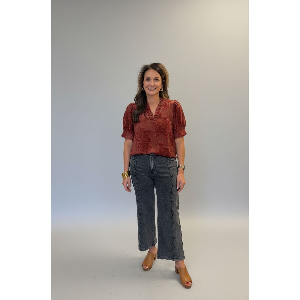 A woman stands indoors wearing a rust-red short-sleeve top with a textured pattern and dark gray wide-leg cropped pants. She is positioned next to a white column in a home or boutique interior with beige walls, tiled floors, and furniture in the background.
