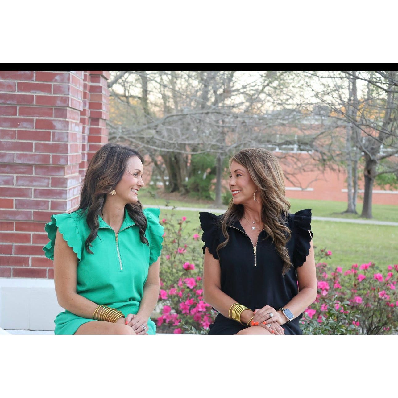Two women sitting outdoors with a brick building and greenery in the background