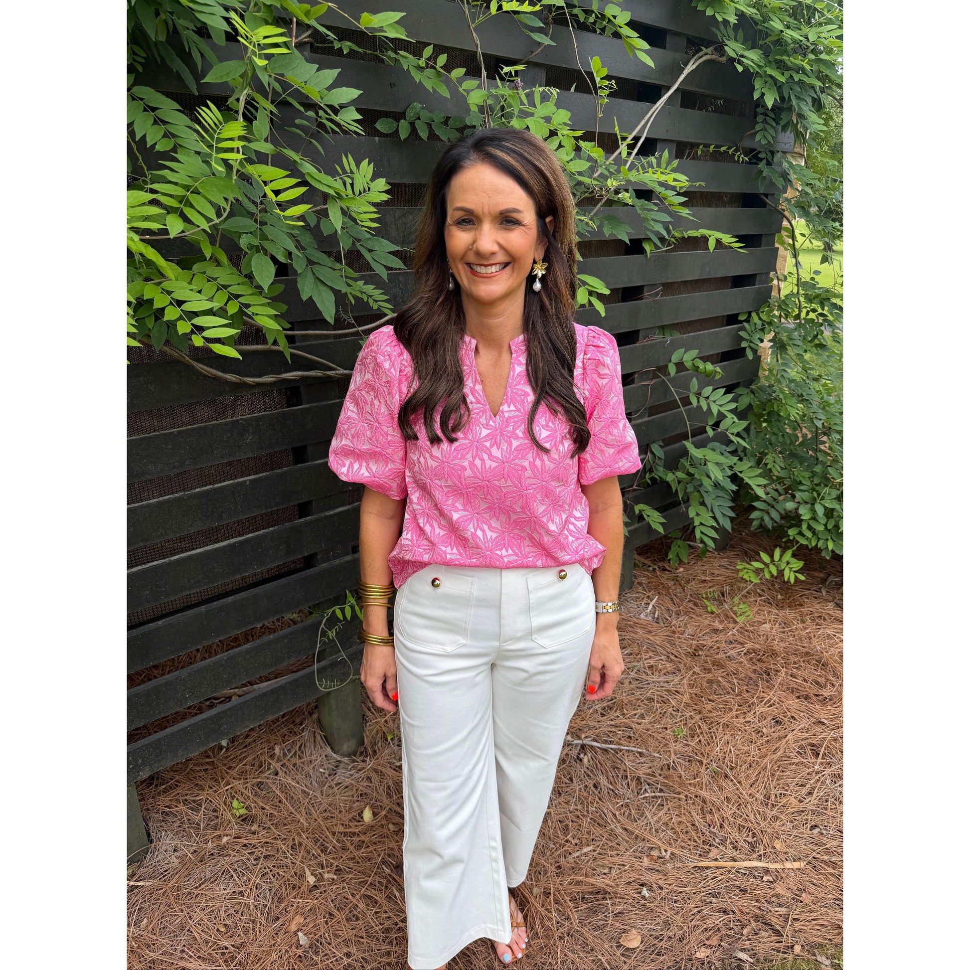 Woman in a pink top and white pants standing outdoors with greenery around