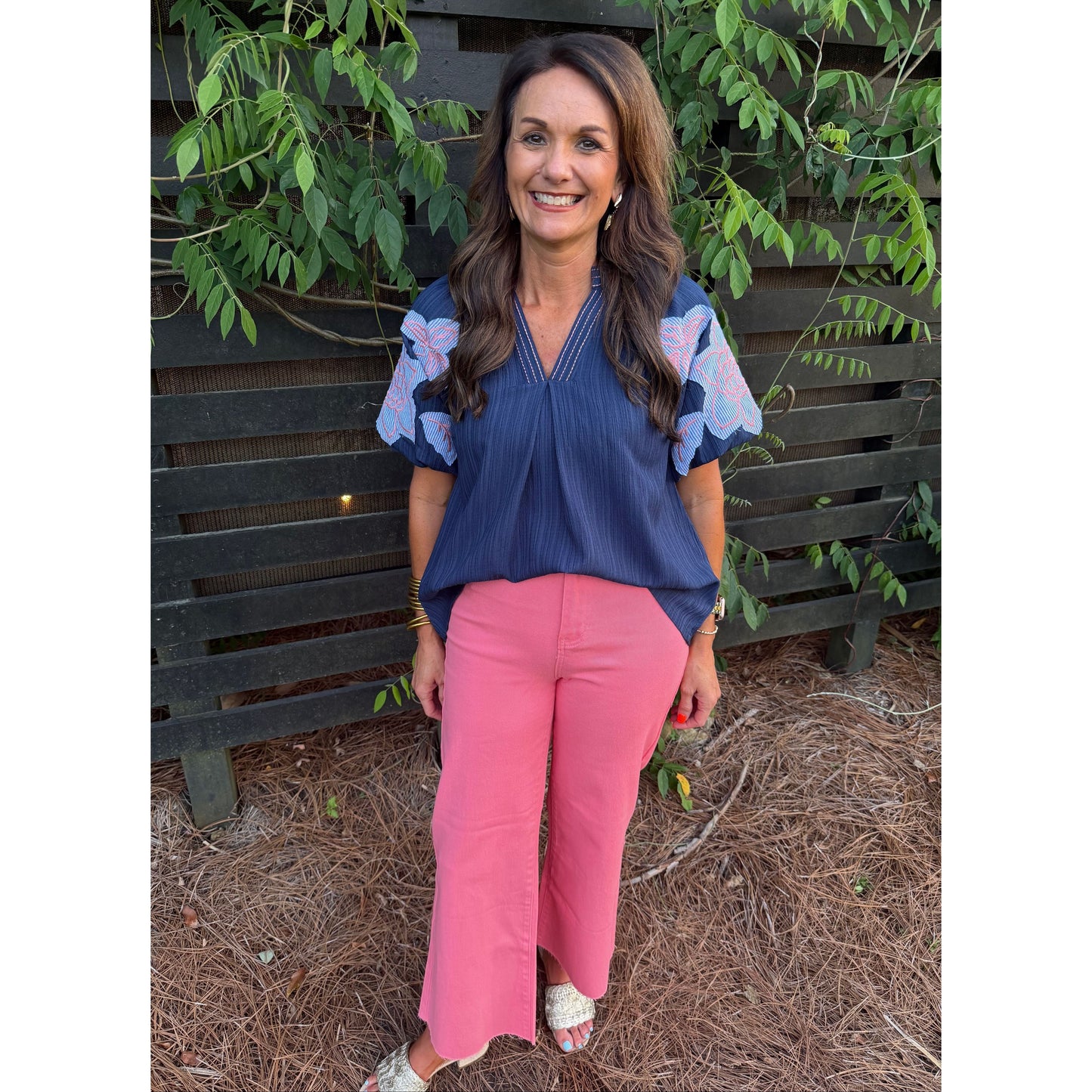 Woman wearing a blue floral blouse and pink pants standing in front of a wooden fence with greenery.