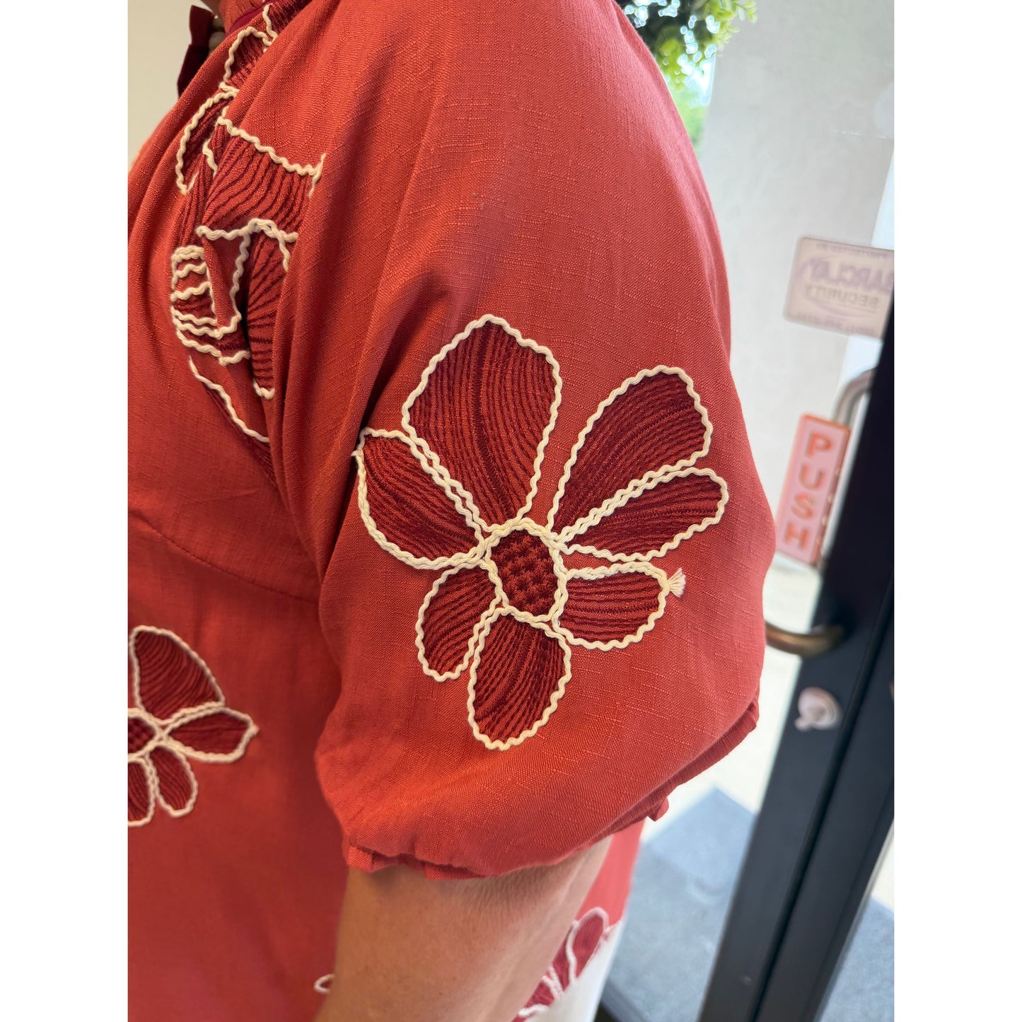 Close-up of a terracotta red short-sleeve shirt with cream-colored floral embroidery on the sleeve, worn by a person standing near a glass door.