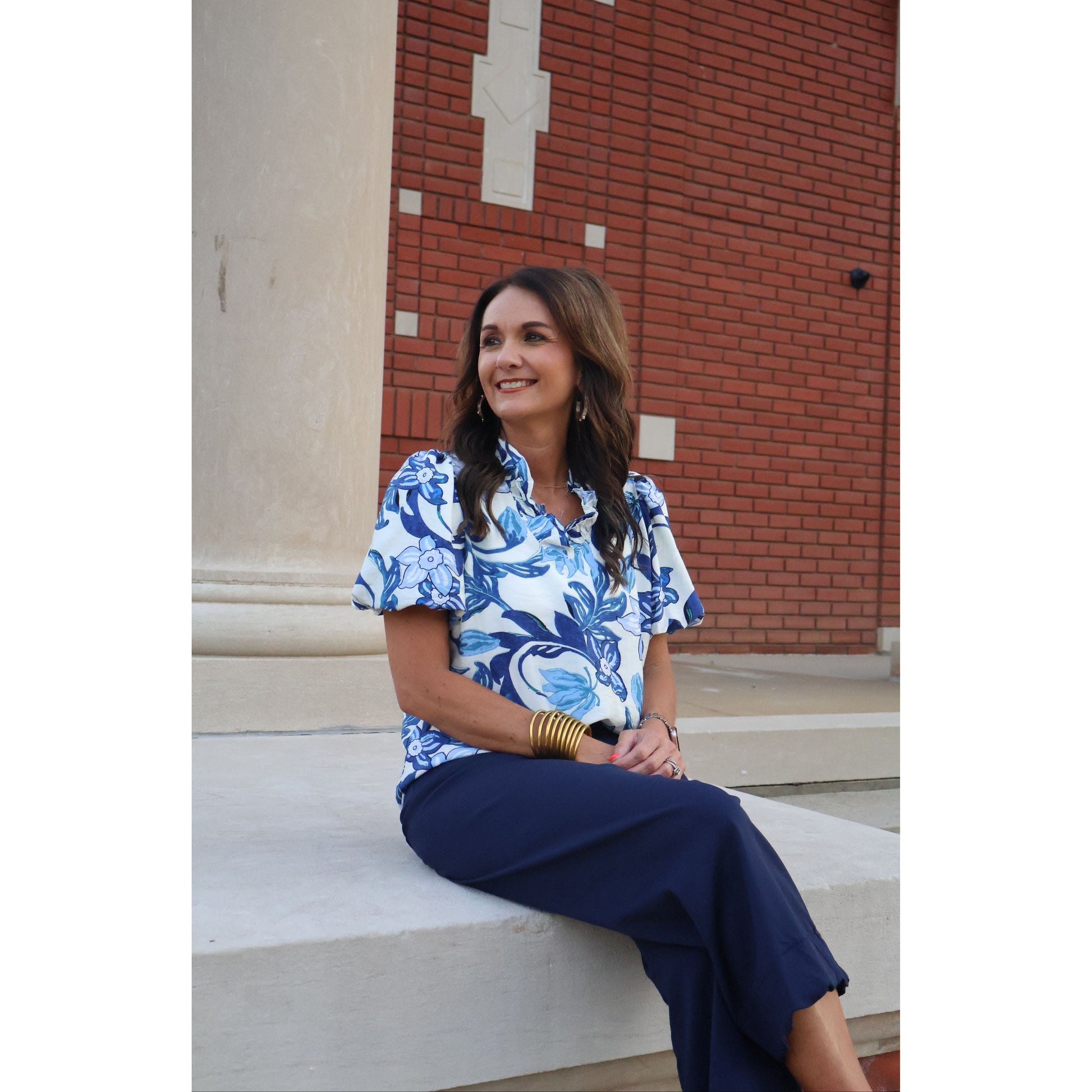 Woman in a blue floral dress standing against a building with a red brick wall.