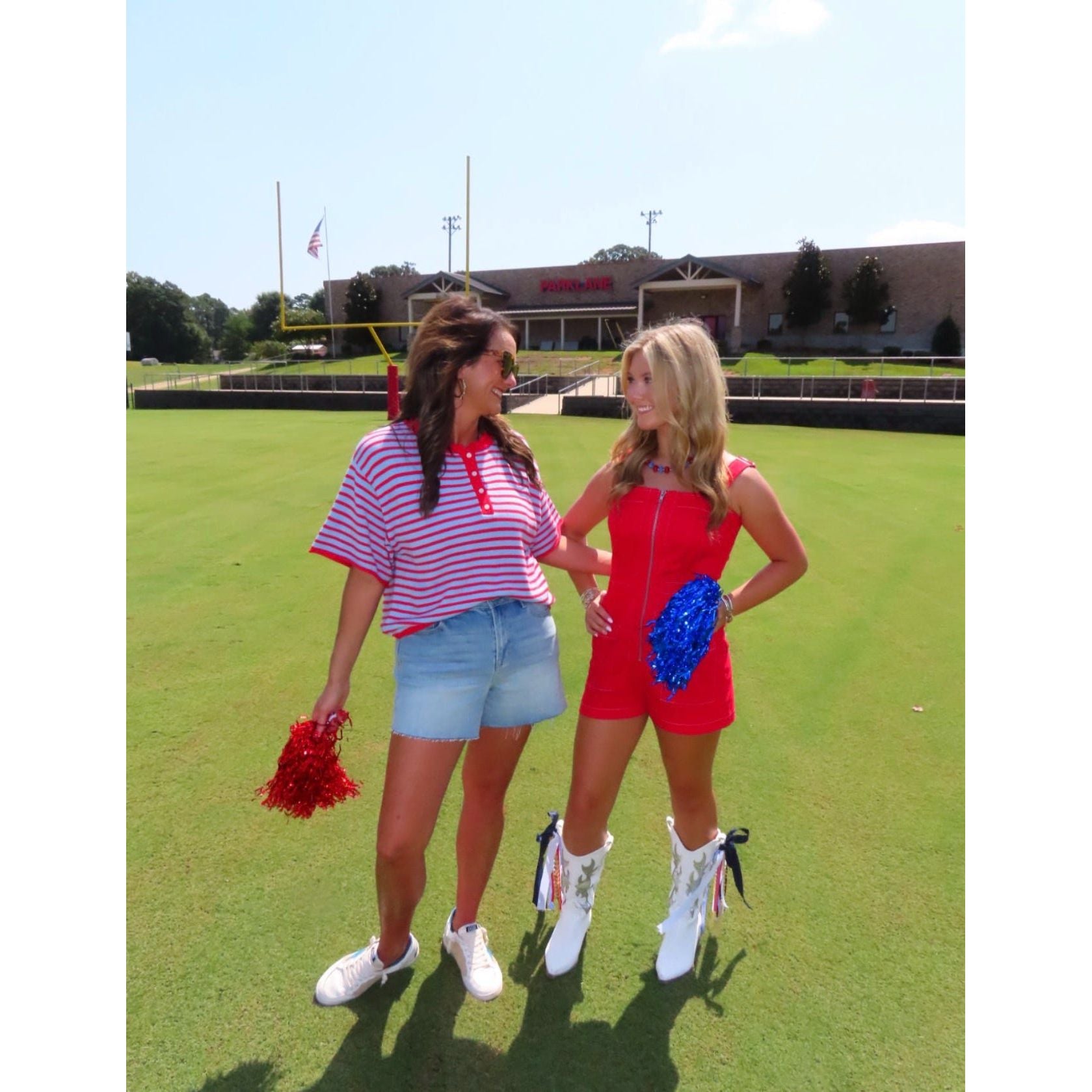 Two women standing on a green grass field wearing cheerleader outfits, one in a pink and white striped shirt with denim shorts holding a red pom-pom, and the other in a red romper with white boots holding a blue pom-pom, with a stadium building in the background.