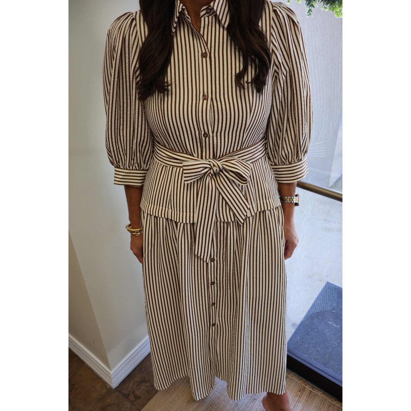 A woman wearing a beige and brown vertically striped midi shirt dress with puff sleeves, a button-down front, and a matching tie belt at the waist.