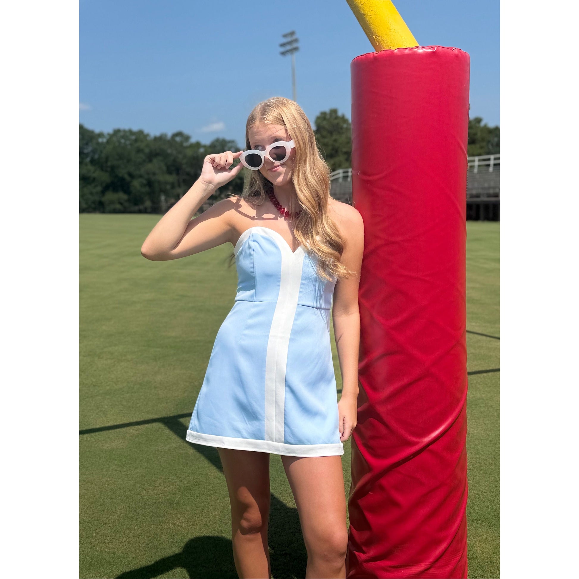 A woman stands on a grassy field wearing a light blue and white strapless mini dress and white sunglasses, posing next to a large red padded goal post.