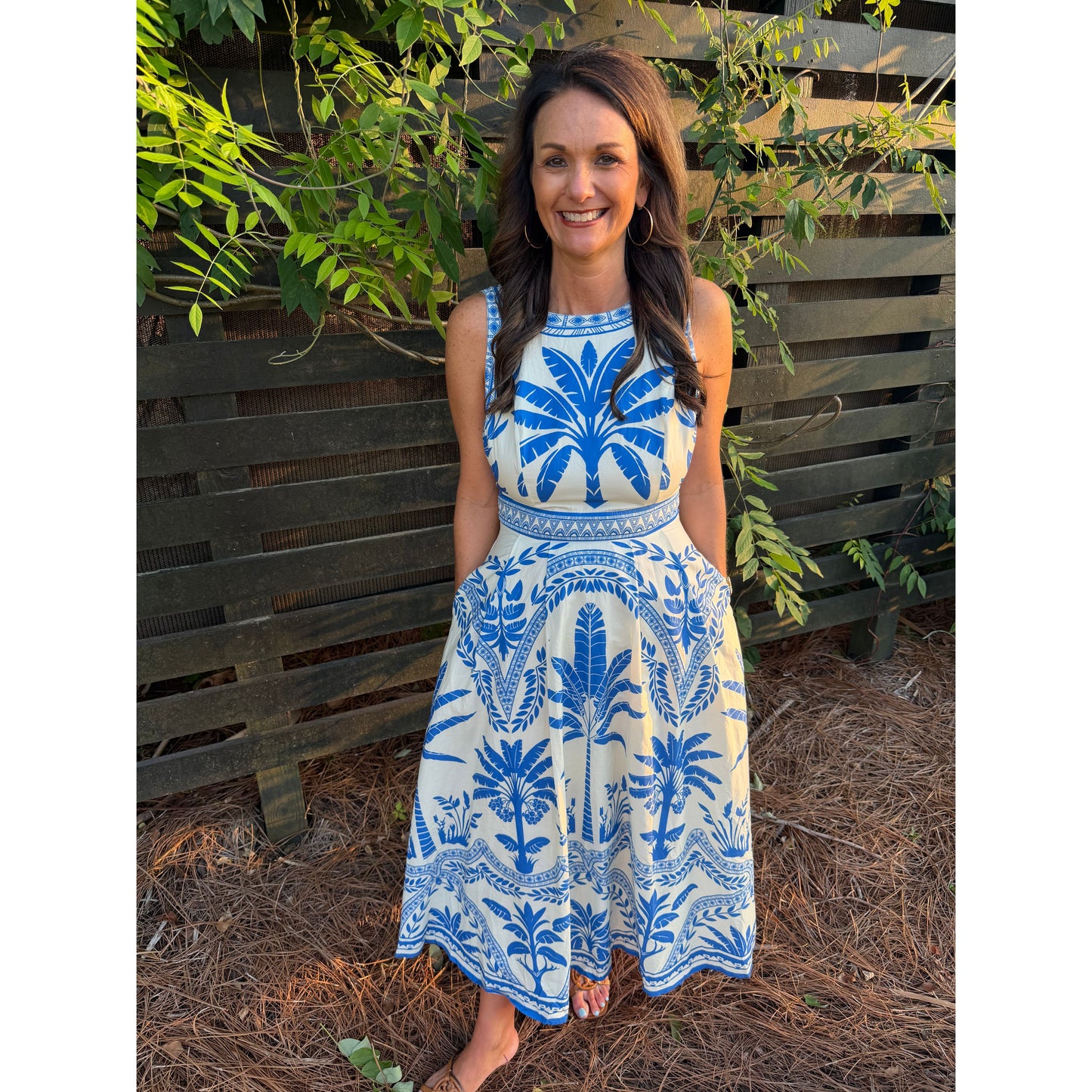 Woman in a blue and white patterned dress standing outdoors with plants around.