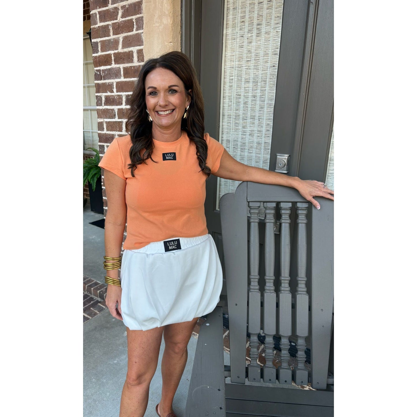 Woman in orange shirt and white skirt standing on a porch