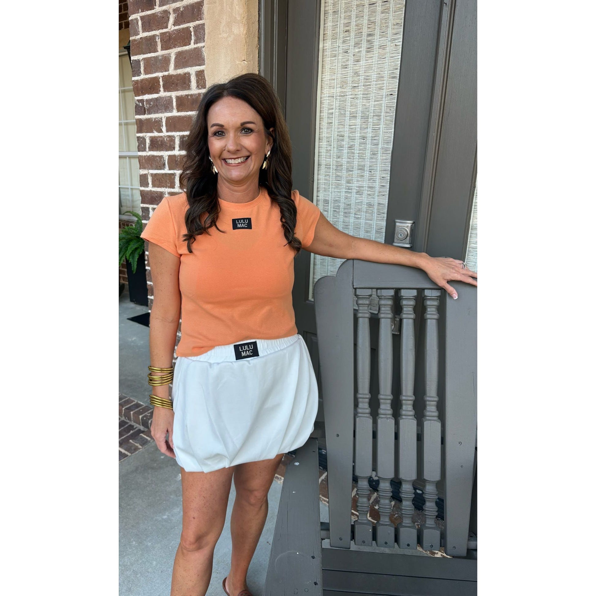 Woman in orange shirt and white skirt standing on a porch