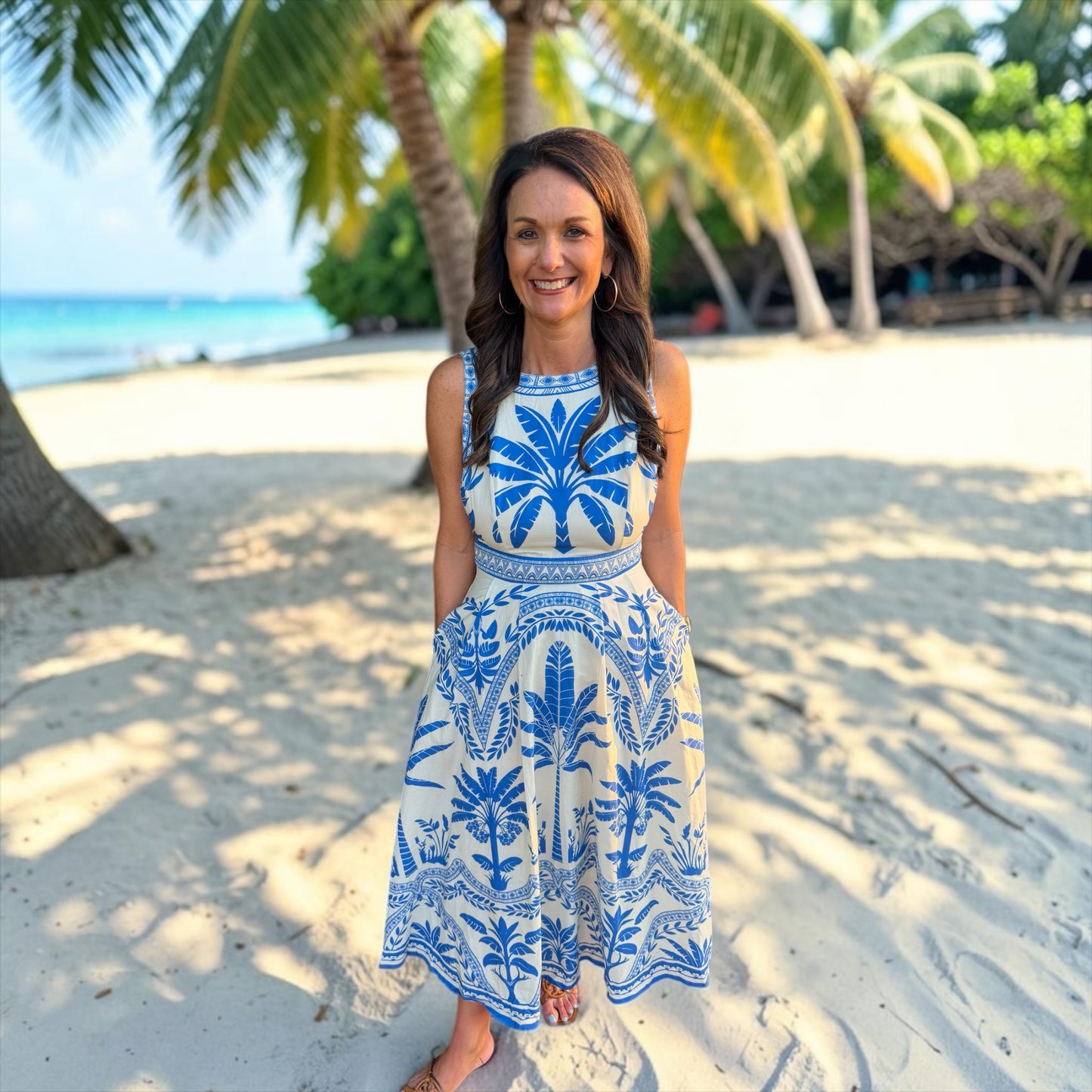 Woman in a blue and white dress standing on a sandy beach with palm trees in the background.