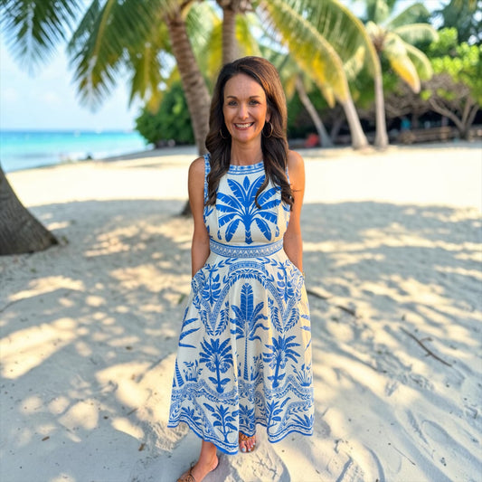 Woman in a blue and white dress standing on a sandy beach with palm trees in the background.