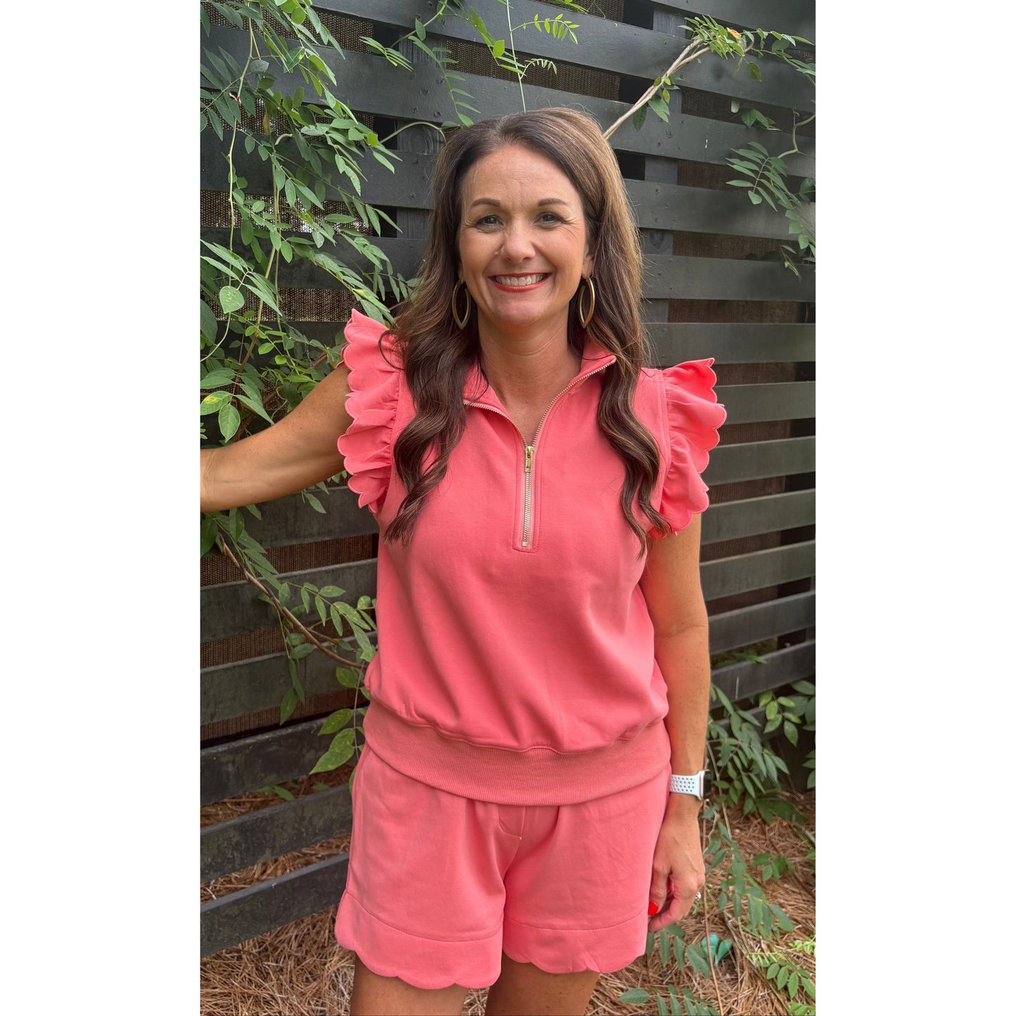 Woman wearing a pink outfit standing against a wooden fence with greenery.