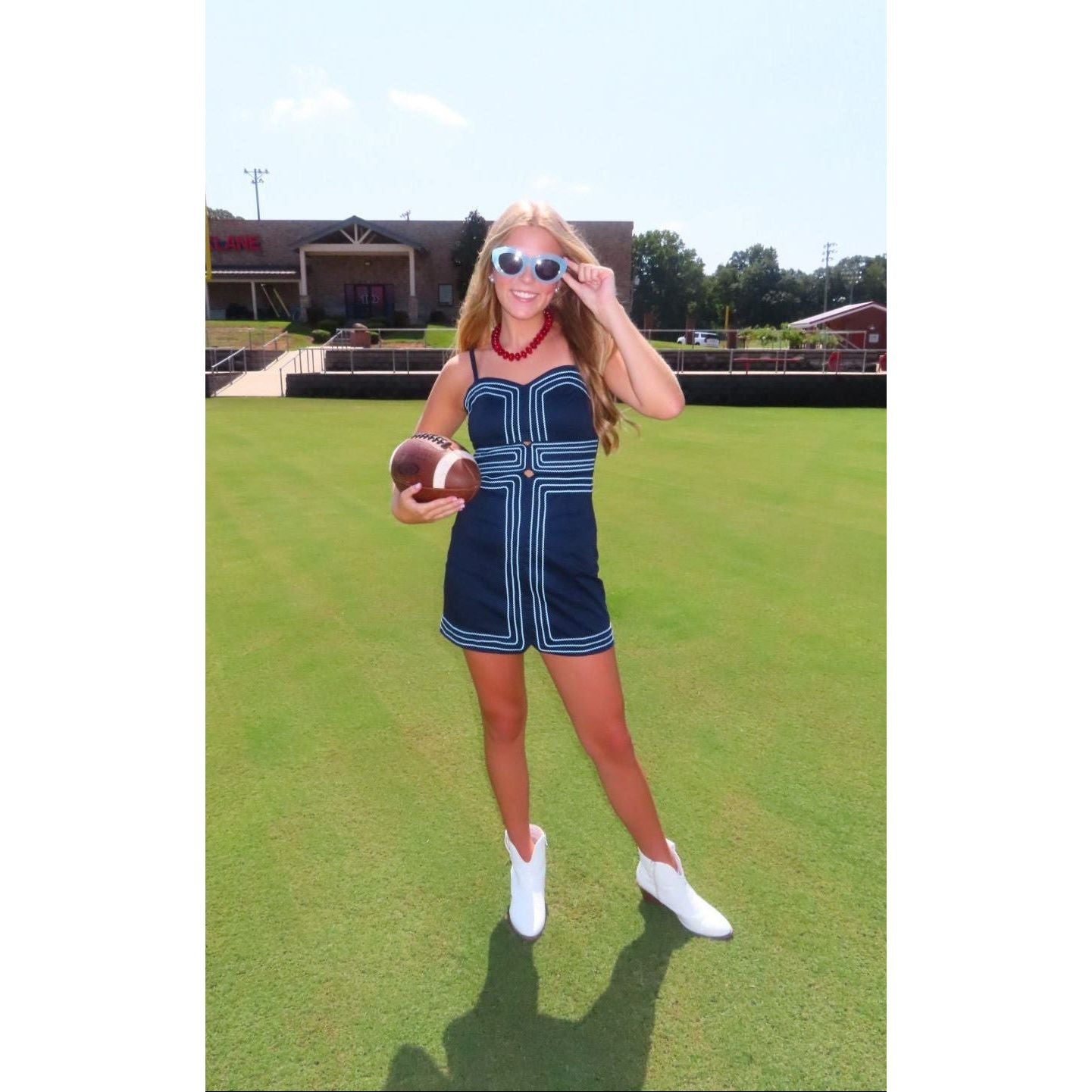 A woman stands on a grassy field wearing a navy blue mini dress with white geometric trim, white cowboy boots, and white sunglasses, holding a football. A building and trees are visible in the background.