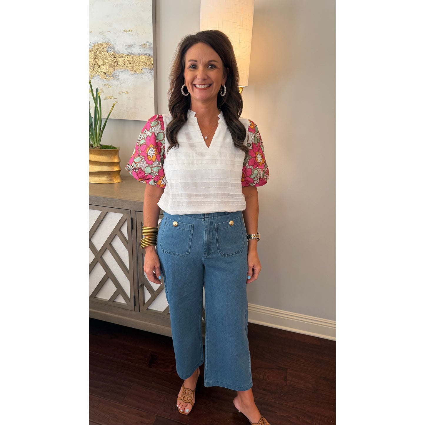 Woman in a white blouse with floral sleeves and blue jeans standing in a room with a lamp and decorative items.