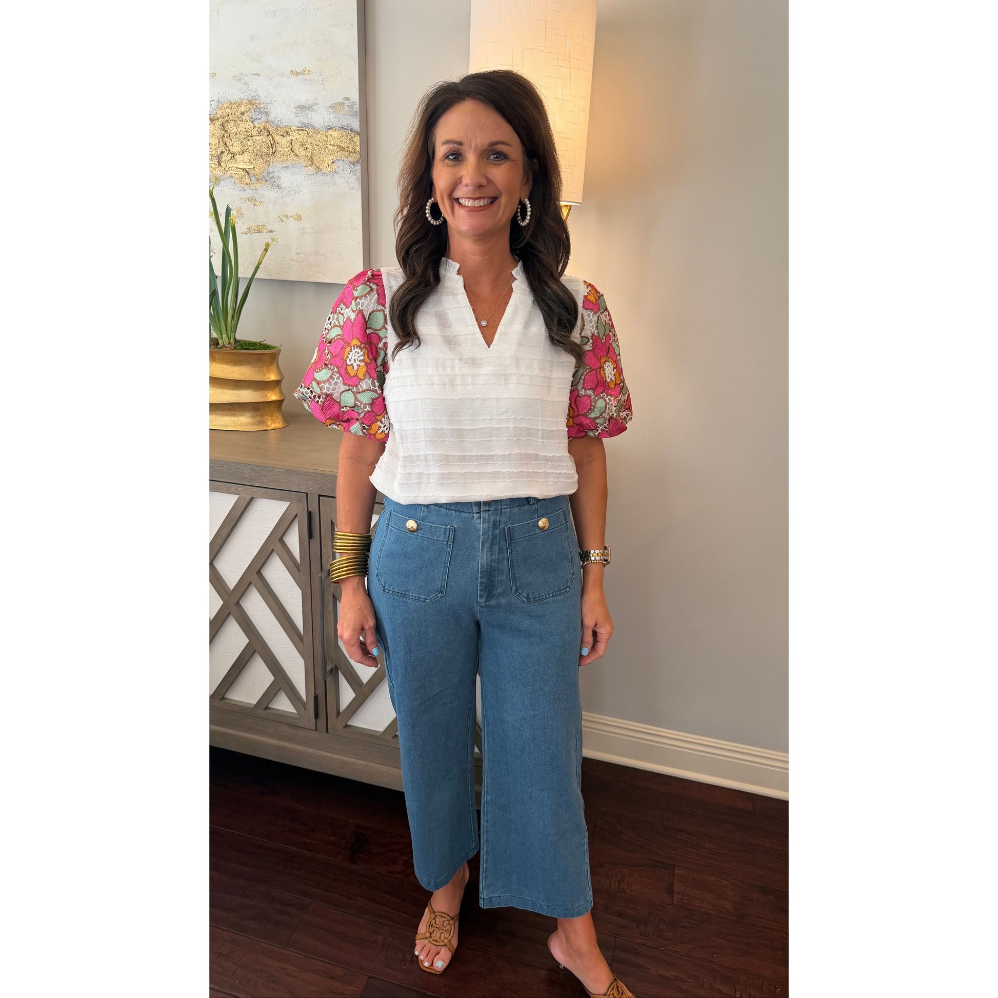 Woman in a white blouse with floral sleeves and blue jeans standing in a room with a lamp and decorative items.