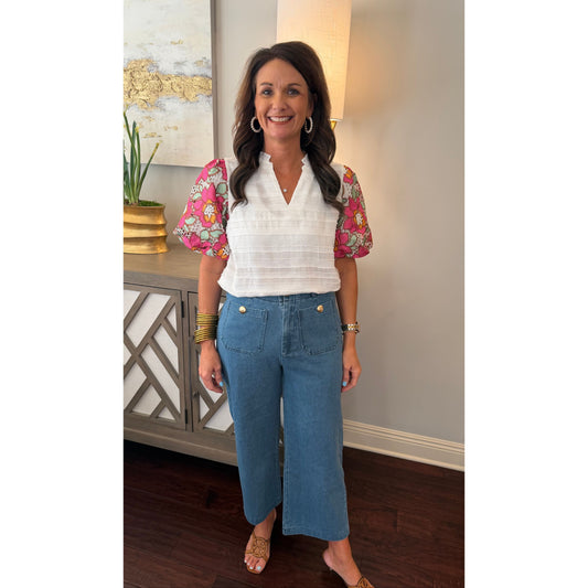 Woman in a white blouse with floral sleeves and blue jeans standing in a room with a lamp and decorative items.