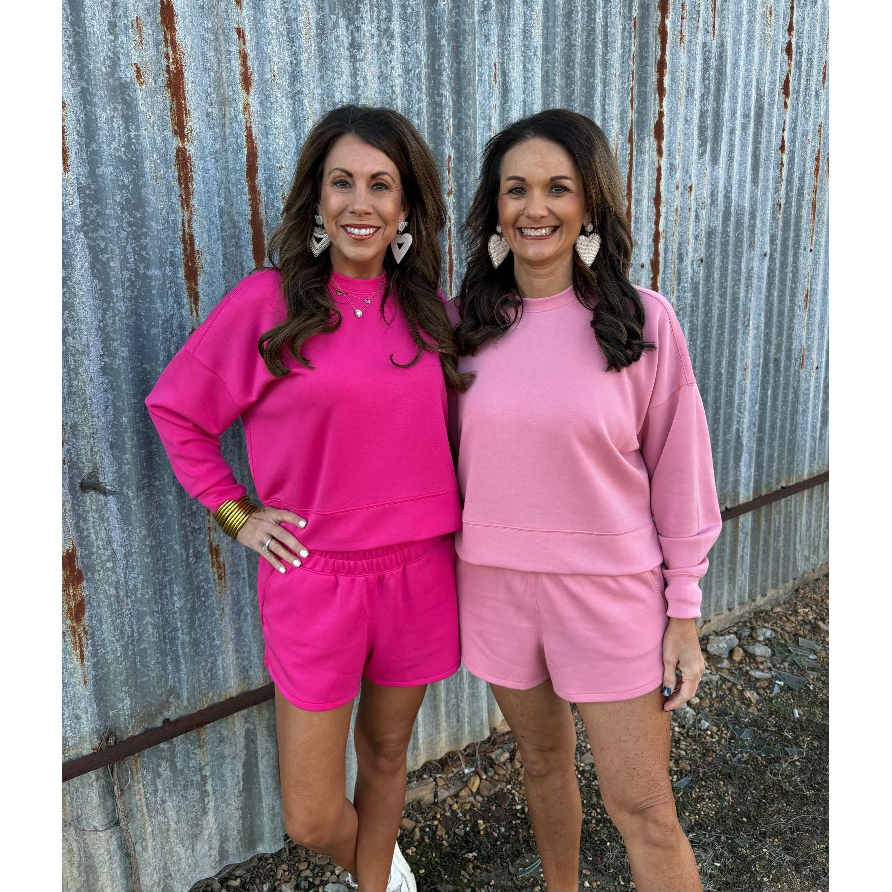 Two women in matching pink outfits standing against a corrugated metal wall.