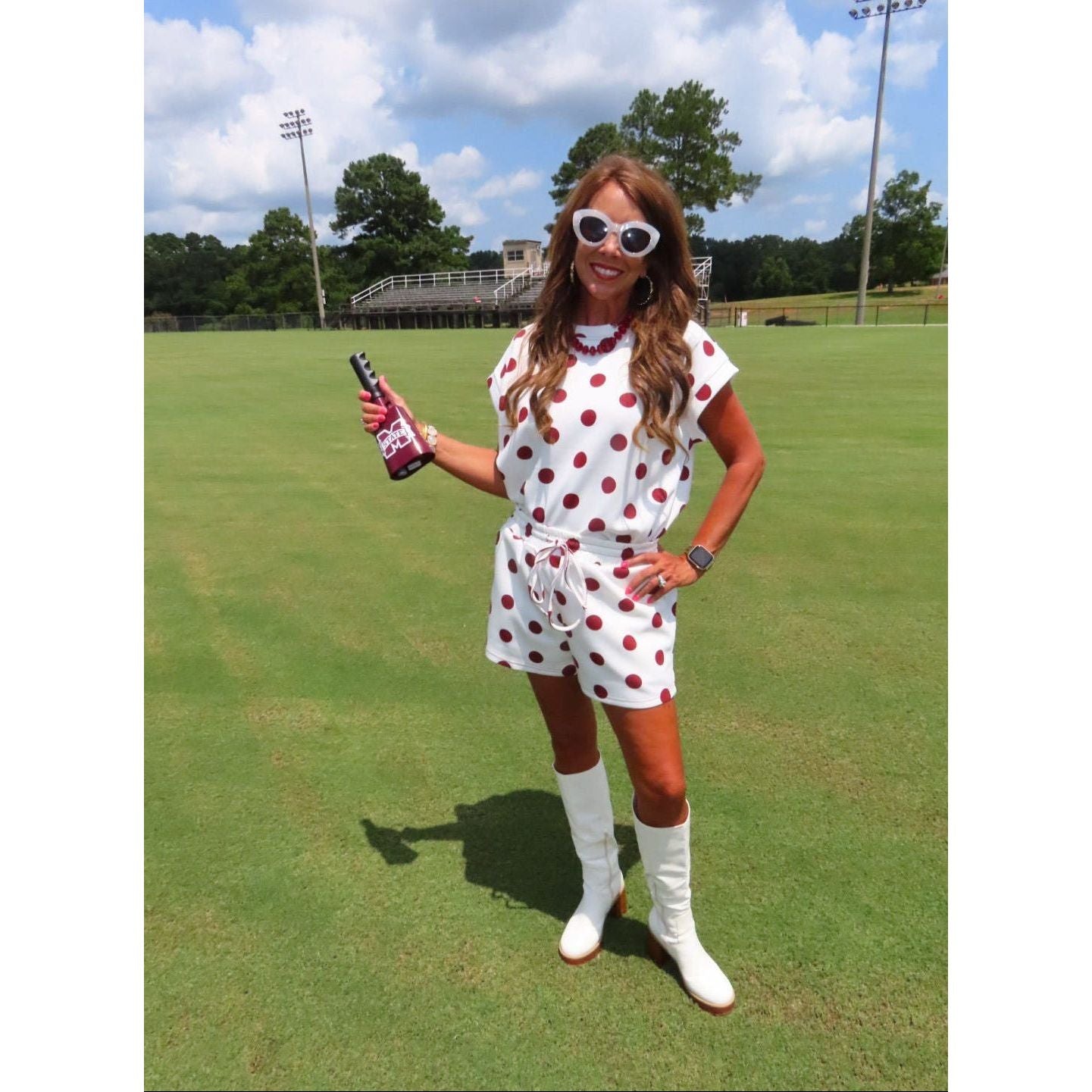 A woman stands on a grassy golf course wearing a white short-sleeve romper with red polka dots, white knee-high socks, and white boots, while holding a bottle.