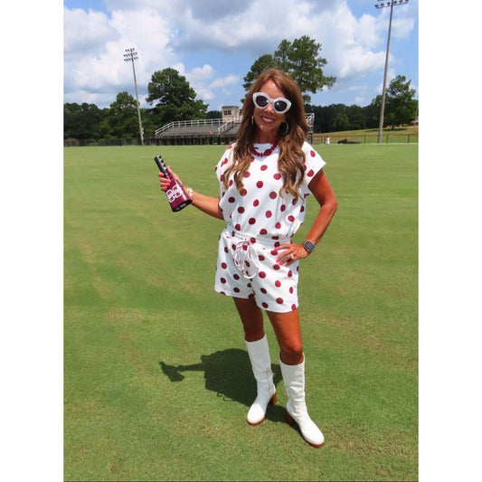 A woman stands on a grassy golf course wearing a white short-sleeve romper with red polka dots, white knee-high socks, and white boots, while holding a bottle.