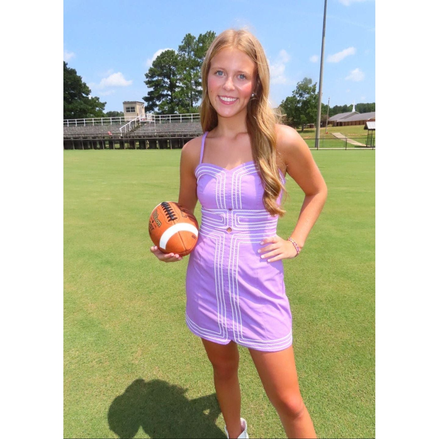 A woman stands on a grassy field outdoors wearing a lavender sleeveless mini dress with white vertical stripe details and holding an American football.
