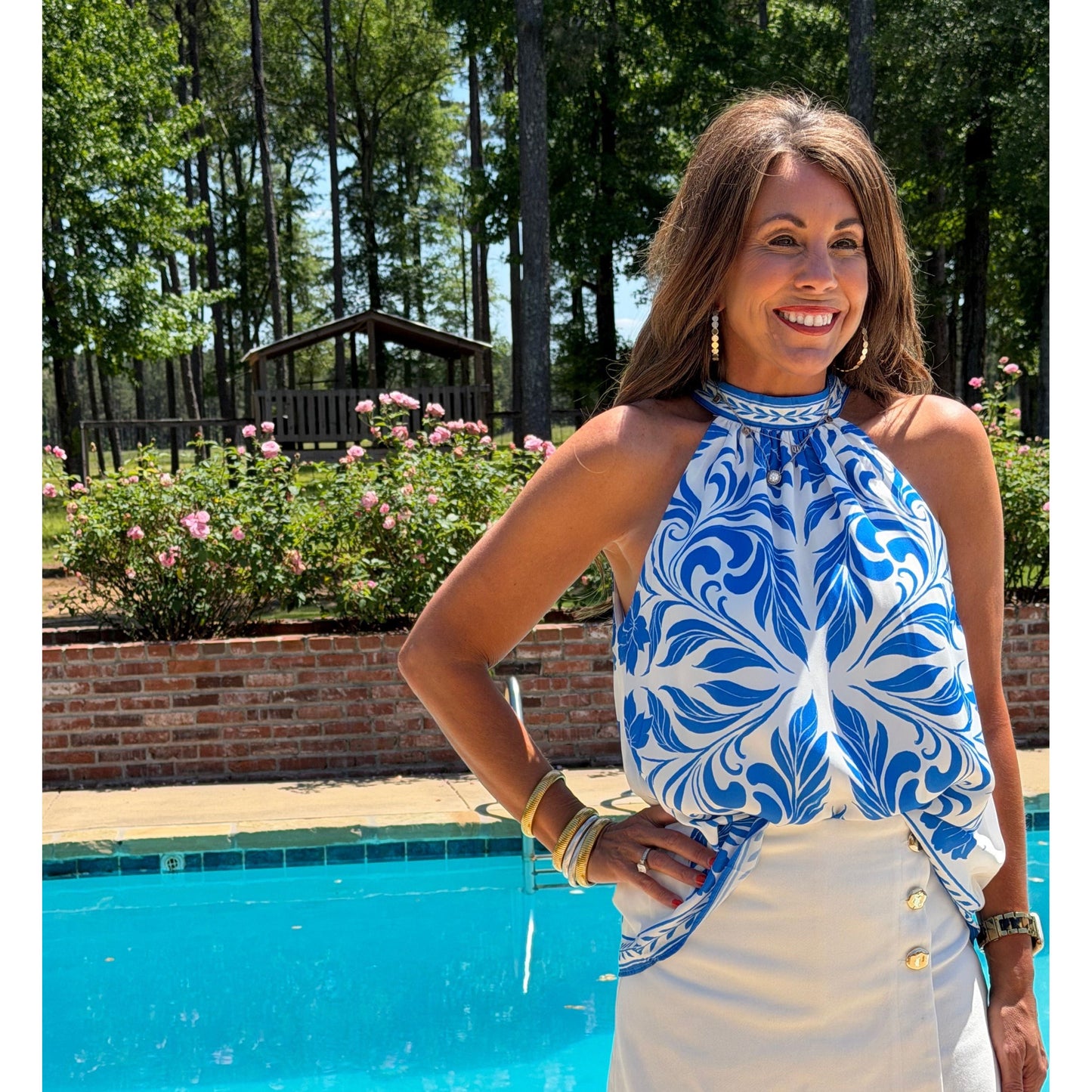 Woman wearing a blue and white patterned top by a pool with trees and flowers in the background