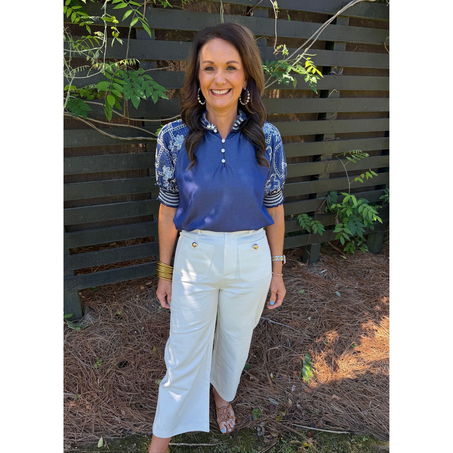 Person wearing a blue top and white pants standing outdoors with plants around