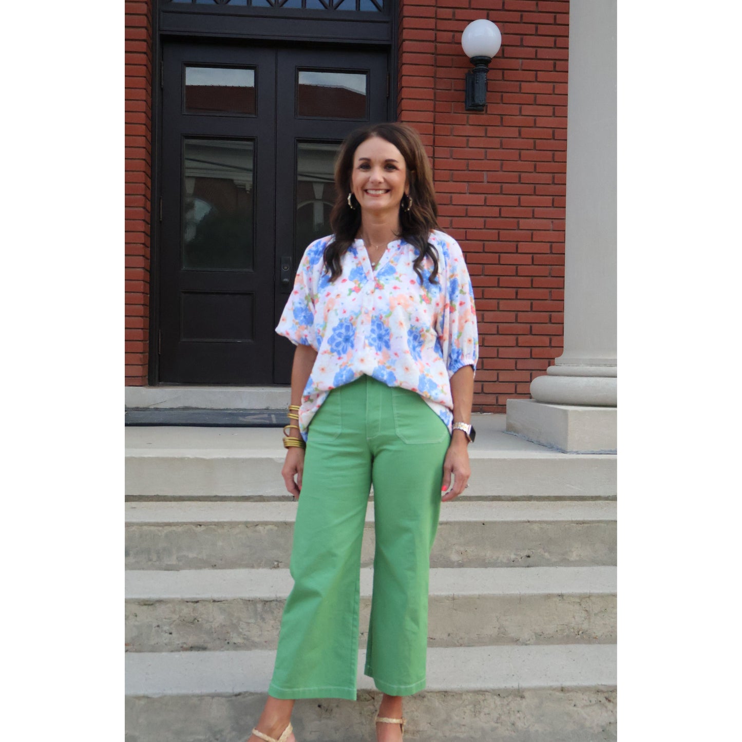 Woman wearing a floral blouse and green pants standing on steps in front of a building.