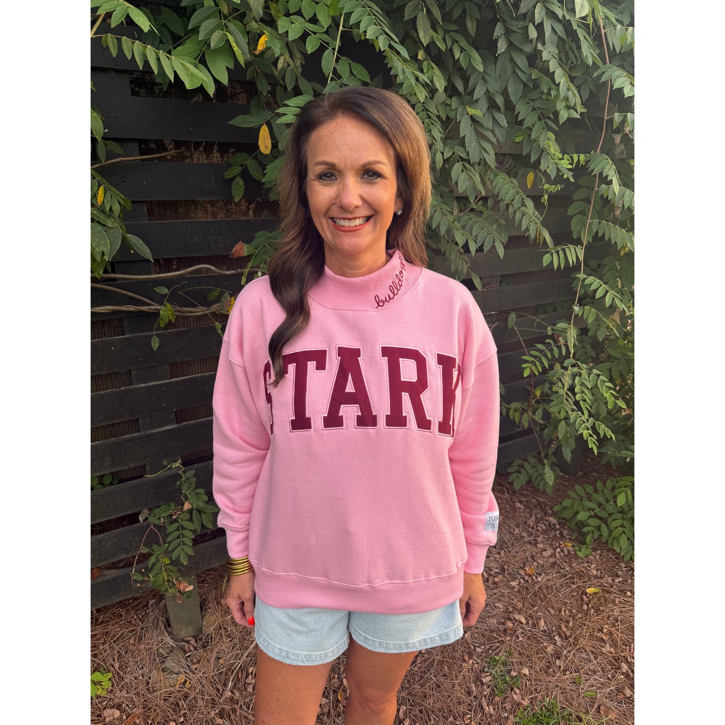 A woman wearing a pink crewneck sweatshirt with 'STARK' printed in maroon block letters across the chest, paired with light denim shorts, standing outdoors in front of a green leafy fence.