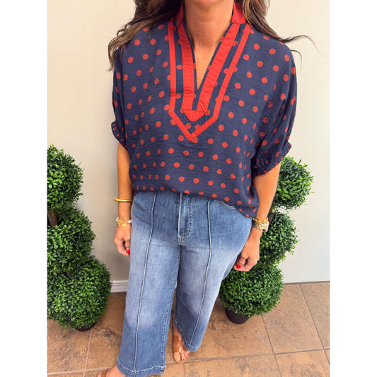 A woman wearing a navy blue blouse with red polka dots and red trim on the V-neck, paired with light-wash blue jeans, standing in front of a beige wall with green potted plants.