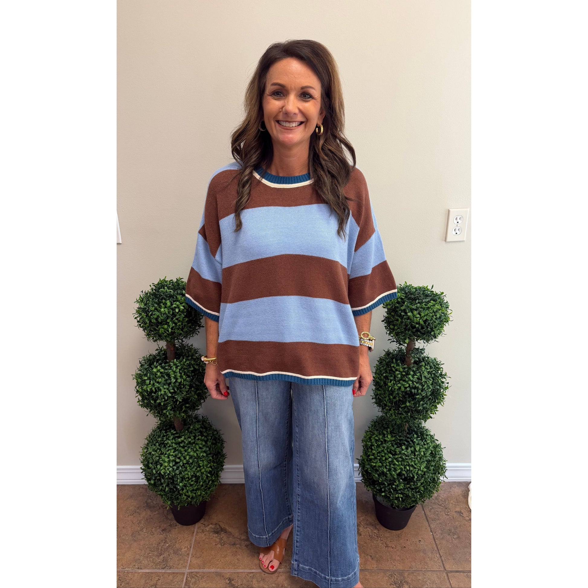 A woman stands indoors wearing a loose knit sweater with wide brown and light blue horizontal stripes and blue jeans. She is positioned between two small potted topiary trees against a plain beige wall.