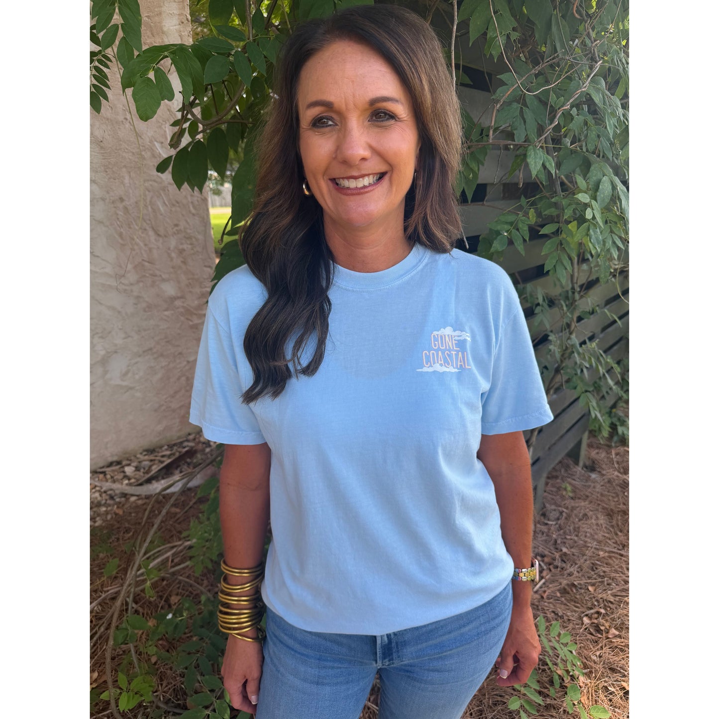 Woman wearing a light blue t-shirt with a logo, standing outdoors among plants.