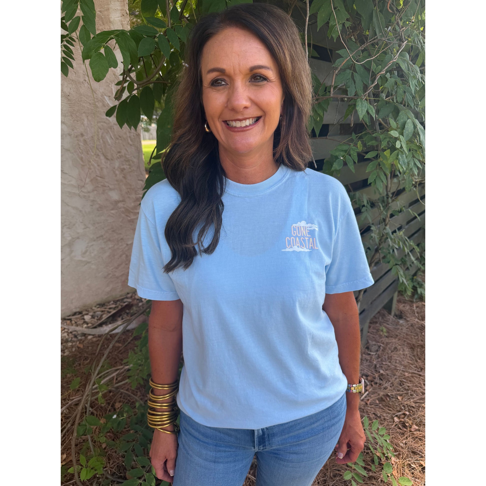 Woman wearing a light blue t-shirt with a logo, standing outdoors among plants.