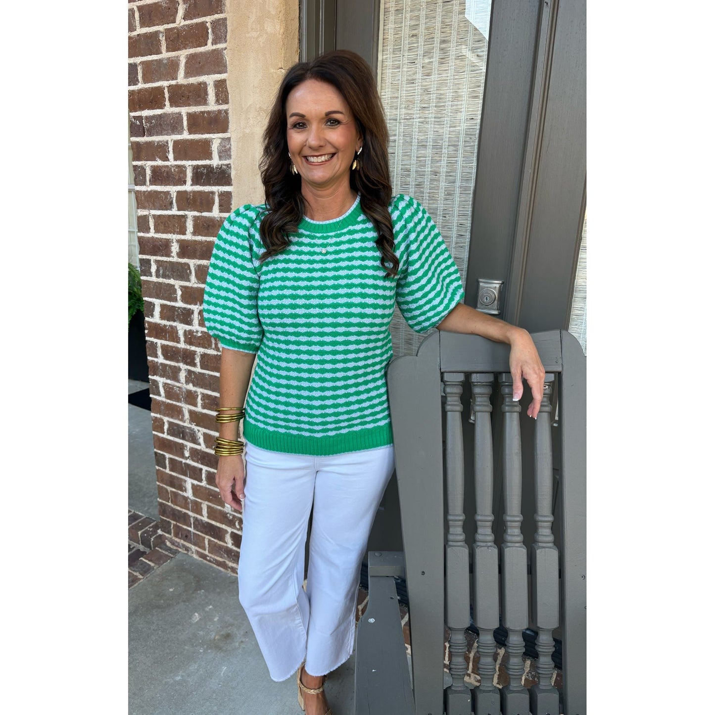 Woman in a green and white checkered shirt and white pants standing on a porch.