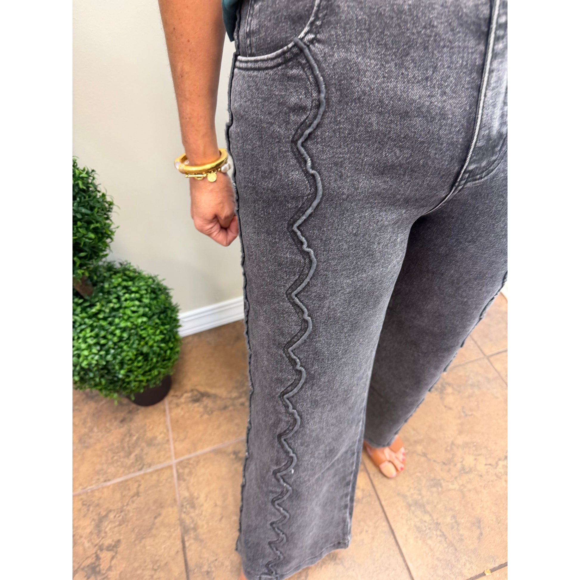 Close-up of a person wearing dark gray washed denim jeans with a distinctive wavy seam detail running down the side of the leg, standing on a tiled floor.