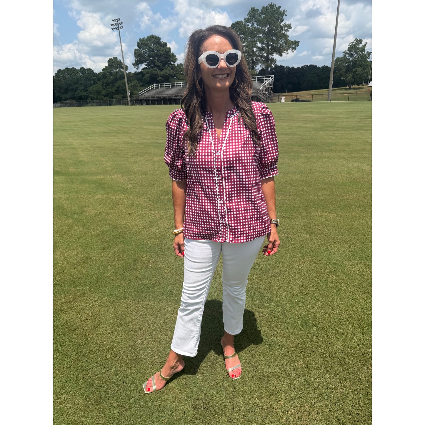 A woman stands on a green grass field wearing a pink and white gingham short-sleeve button-up shirt, white pants, and white-framed sunglasses.