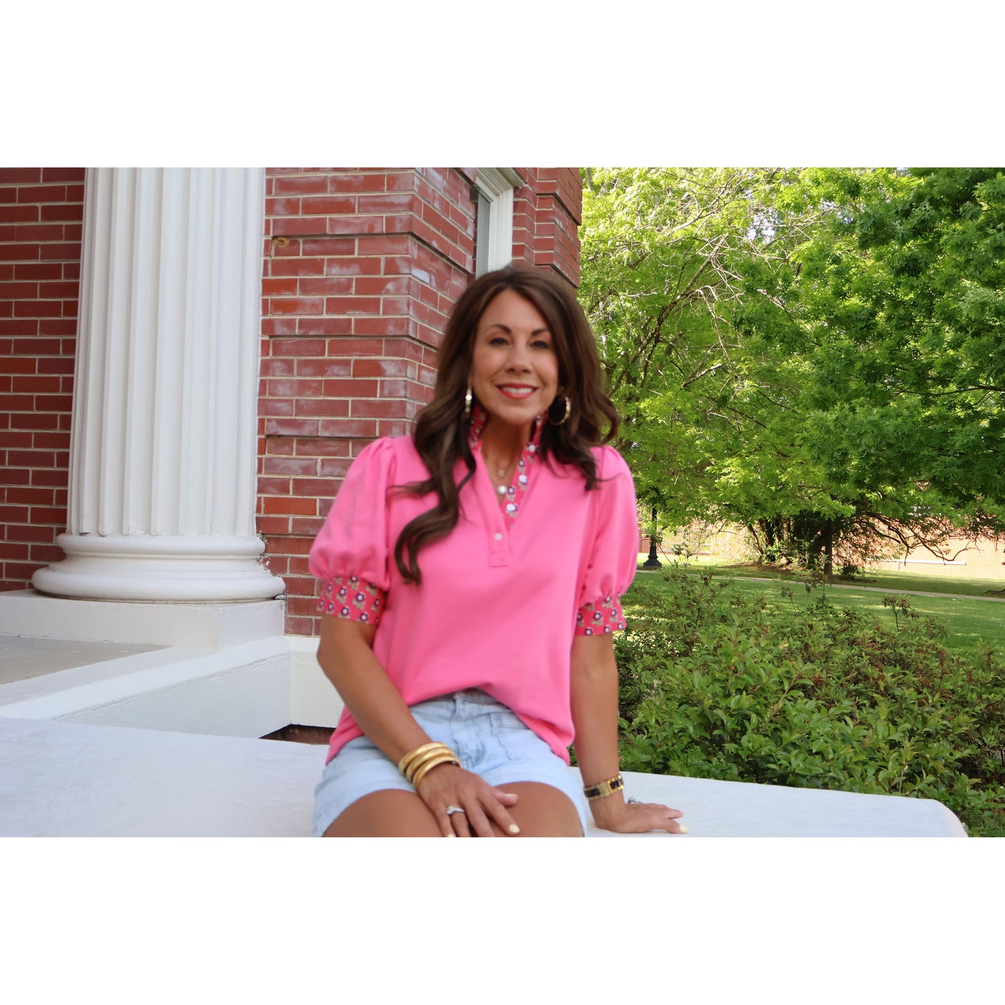Woman in a pink shirt sitting outdoors with a brick building and greenery in the background