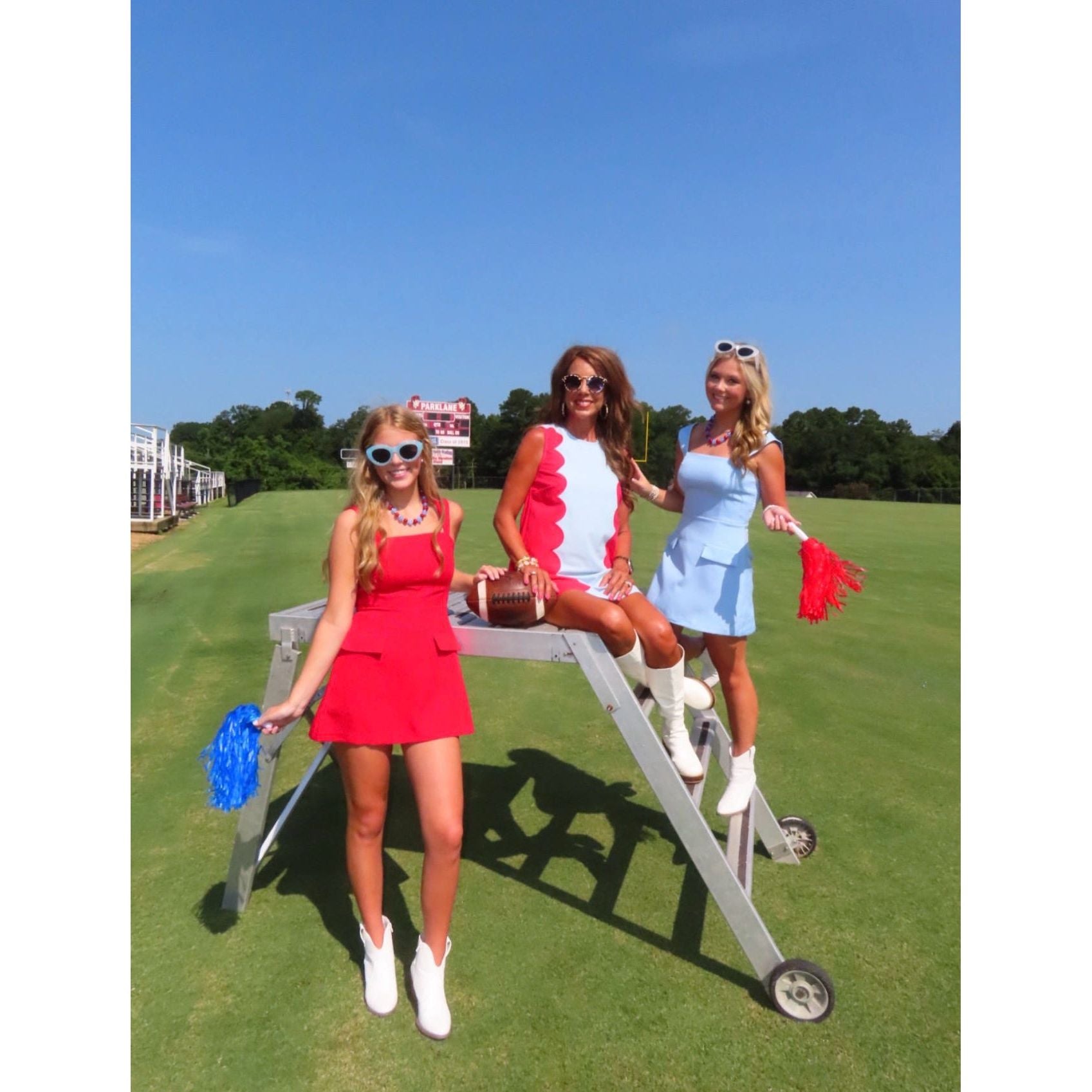 Three women posing on a golf course lift wearing coordinated summer outfits in red, white, and light blue, holding red and blue pom-poms against a blue sky background.