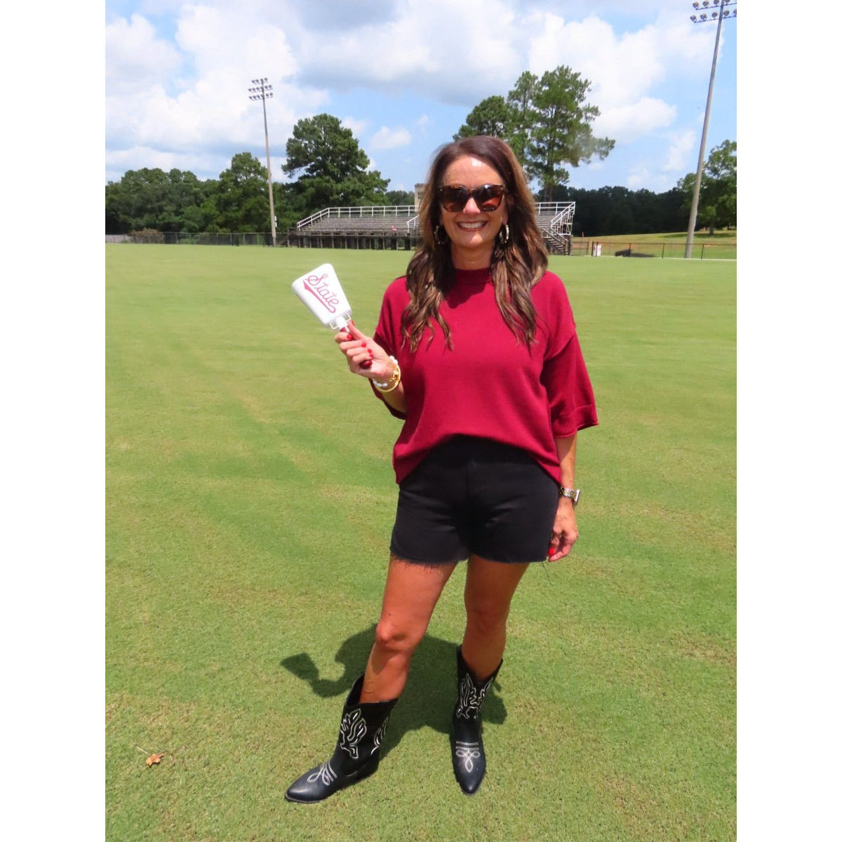 A woman stands on a grassy field outdoors, wearing a bright red short-sleeve top, black shorts, and black cowboy boots with white embroidery. She is holding a white product card in one hand.