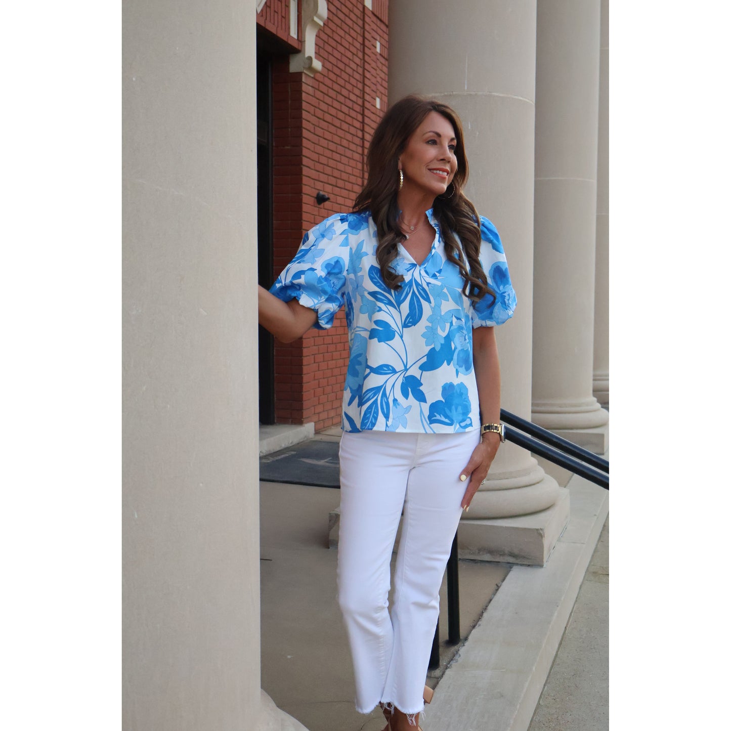 Woman wearing a blue floral dress and white pants standing in a room with a staircase.