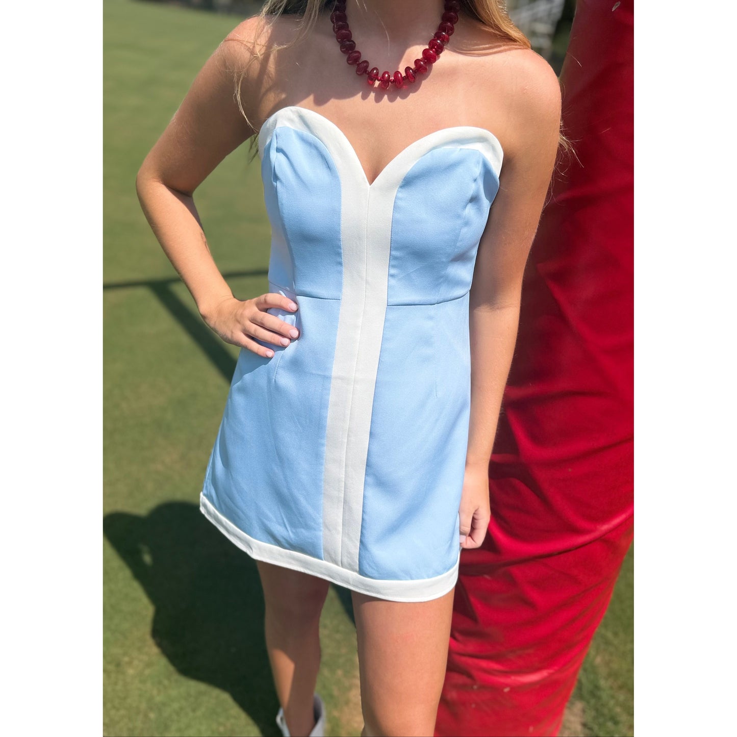 A woman wearing a strapless light blue and white mini dress, standing outdoors on a grassy field next to a red structure.