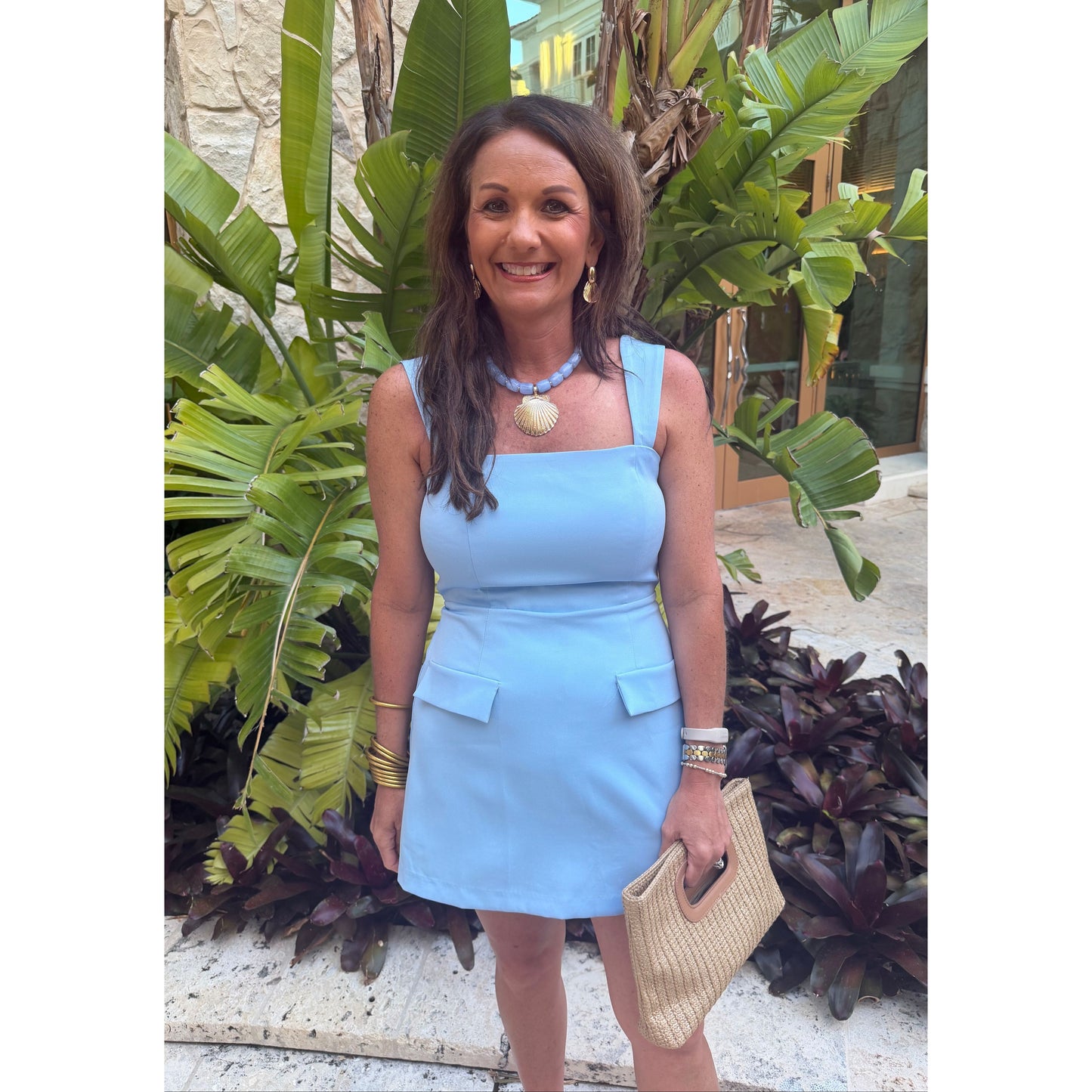 A woman wearing a light blue sleeveless mini dress with a square neckline, structured shoulders, and flap pockets, standing outdoors in front of tropical green plants and holding a woven beige handbag.