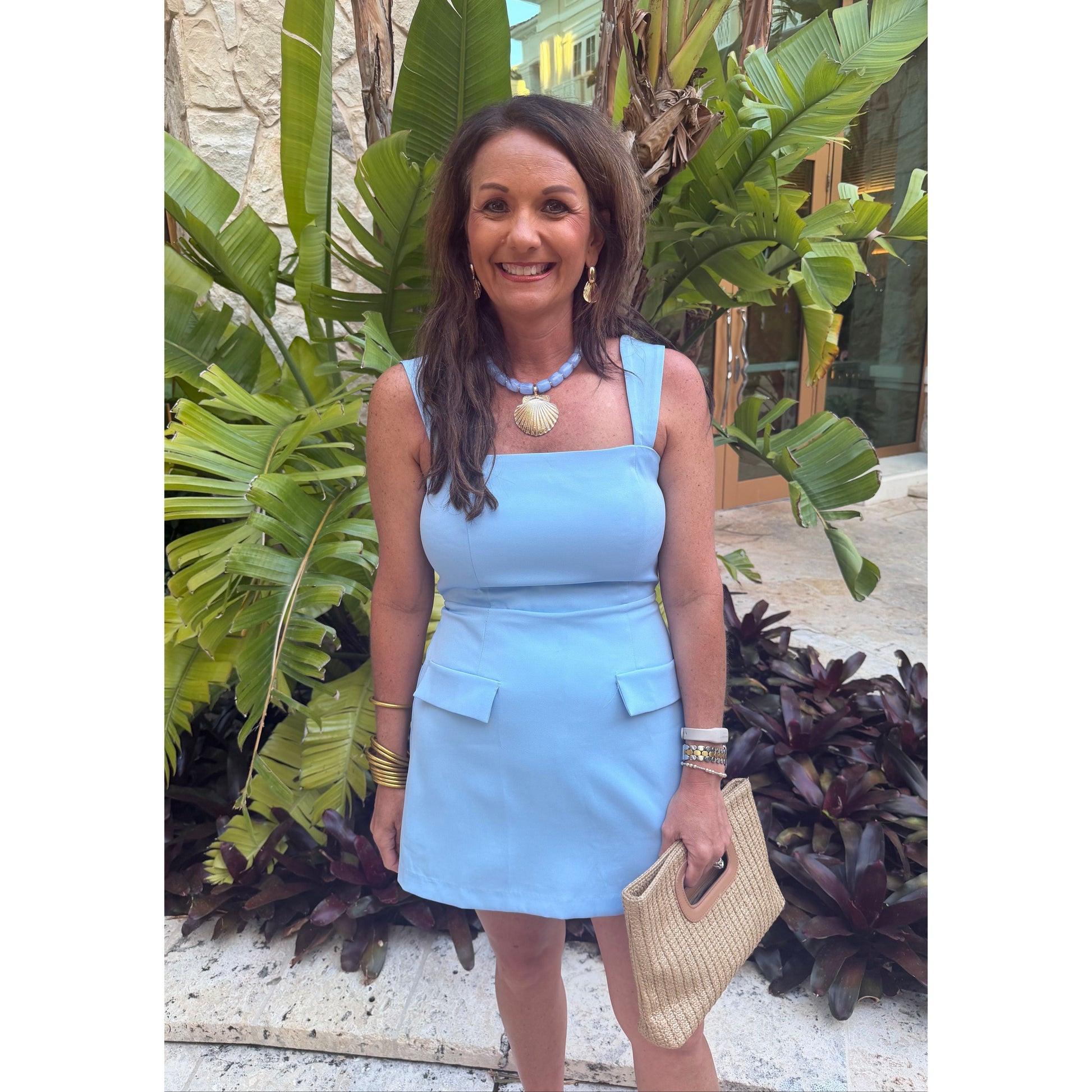 A woman wearing a light blue sleeveless mini dress with a square neckline, structured shoulders, and flap pockets, standing outdoors in front of tropical green plants and holding a woven beige handbag.