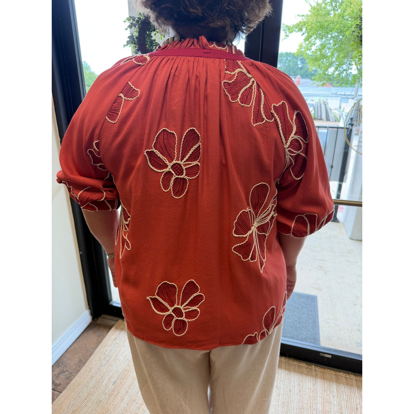 A person viewed from behind wearing a rust-orange short-sleeve blouse with beige floral embroidery and light beige pants, standing indoors near a glass door.