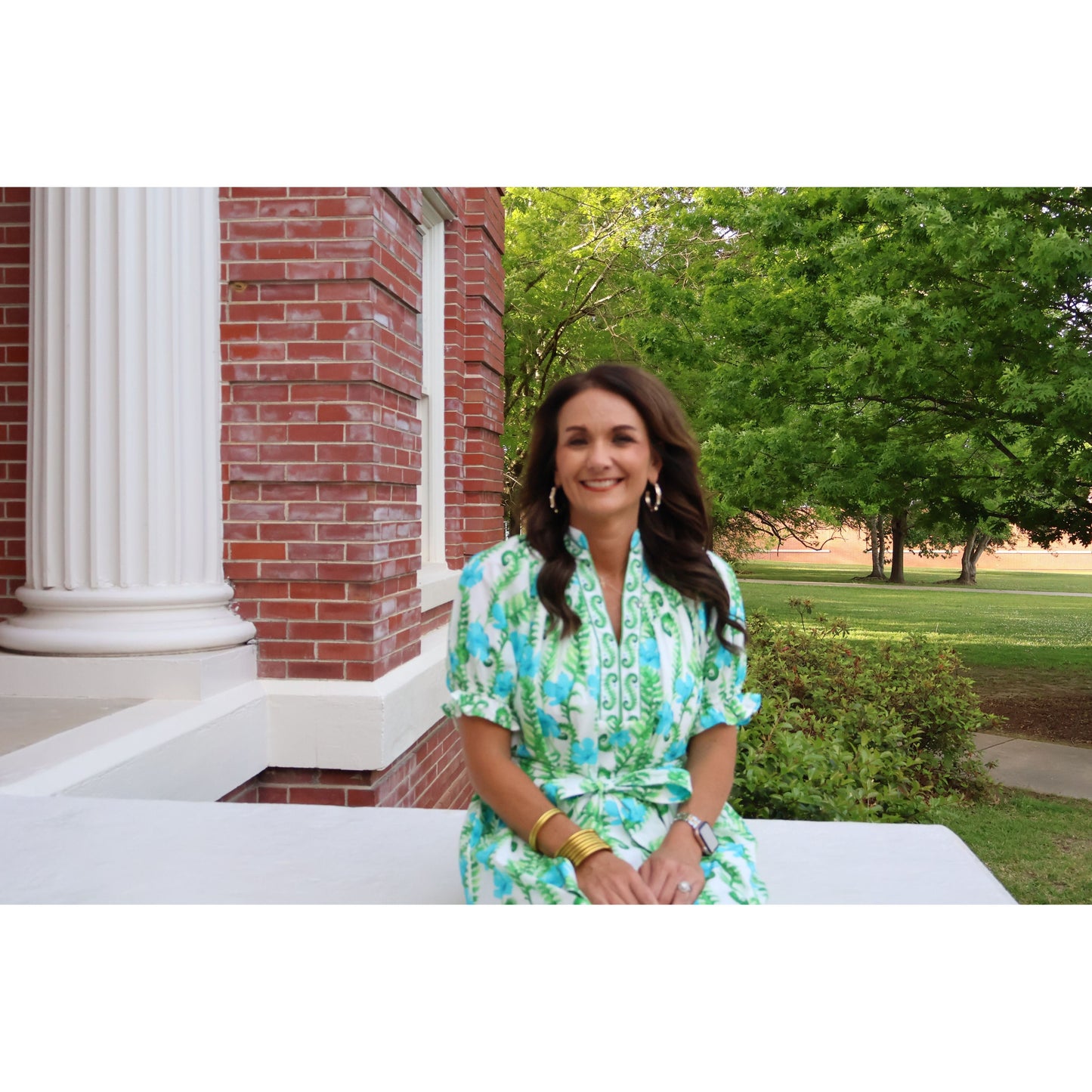 Woman in a green and white dress sitting outdoors near a brick building with trees in the background