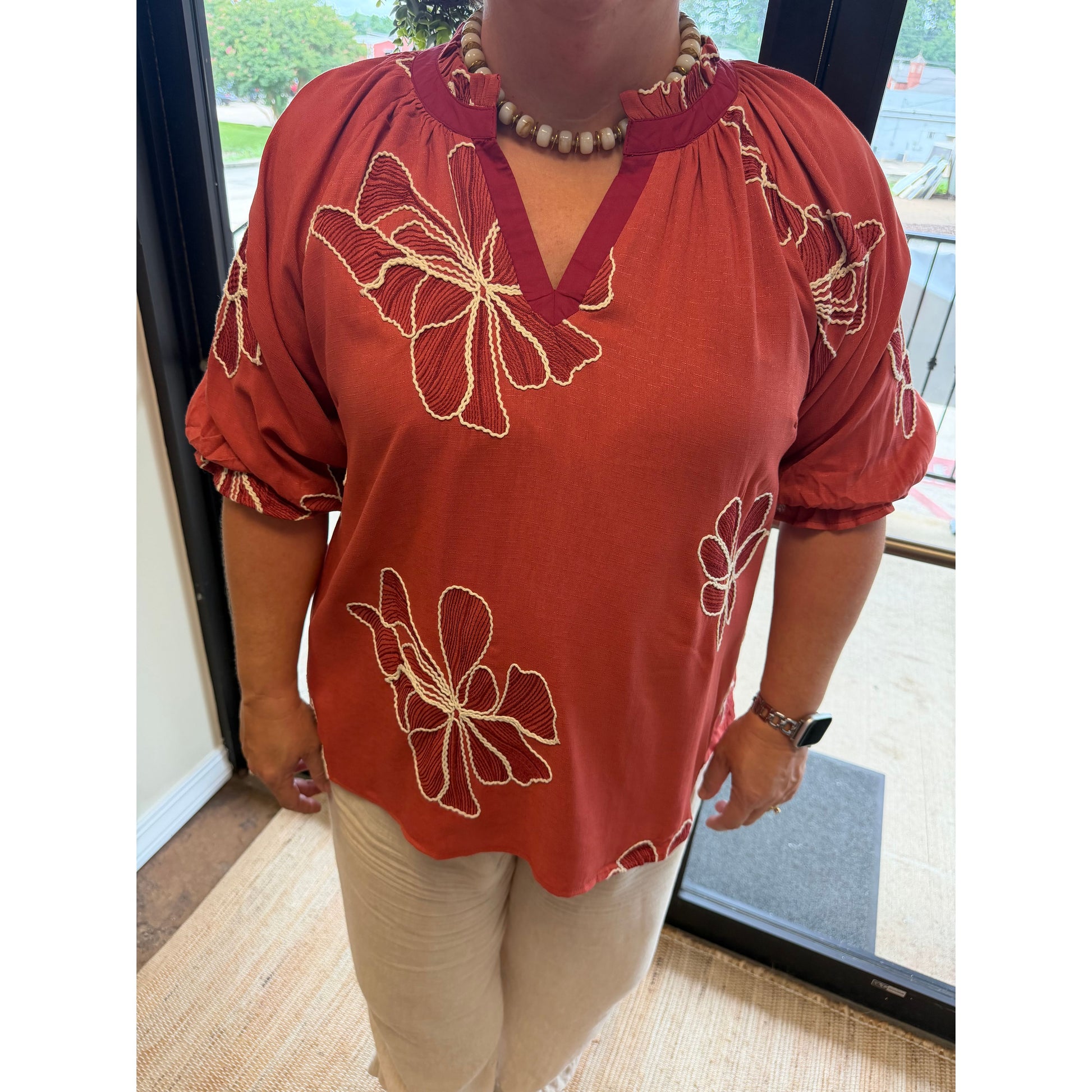A person wearing a rust-orange short-sleeve blouse with beige floral embroidery and a split neckline, paired with light beige pants, standing indoors near a glass door.