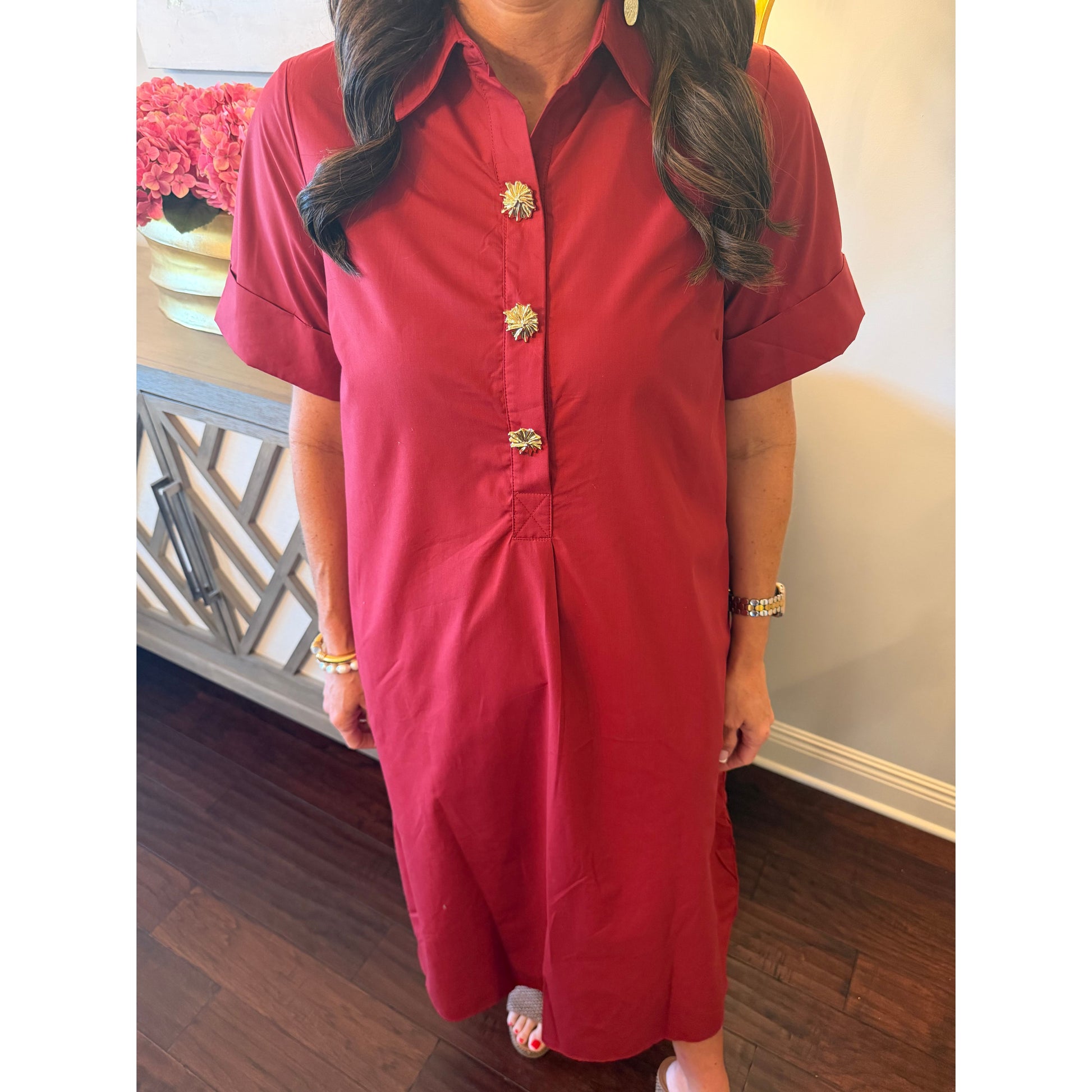 A woman wearing a short-sleeve collared red dress with decorative gold-tone floral buttons down the front, standing indoors on a dark wood floor.
