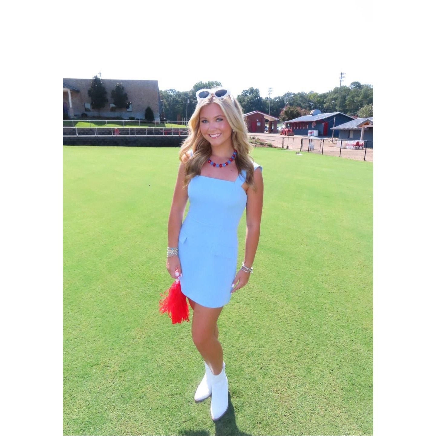 A woman stands on a green grass field wearing a light blue sleeveless mini dress, white ankle boots, and holding a red tassel accessory. She has sunglasses on her head and jewelry on her wrists.