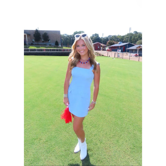 A woman stands on a green grass field wearing a light blue sleeveless mini dress, white ankle boots, and holding a red tassel accessory. She has sunglasses on her head and jewelry on her wrists.
